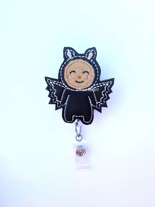 A black felt bat costume badge reel pull with a smiling brown face, white stitched details, and a silver retractable clip on a white background.