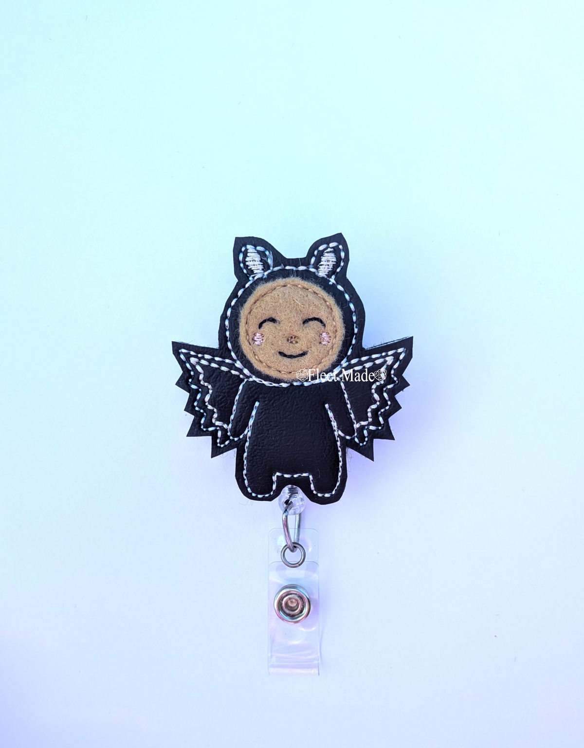 A cute black bat-shaped badge reel holder with a smiling tan face, white stitching, and a clear plastic clip at the bottom, shown on a light blue gradient background.